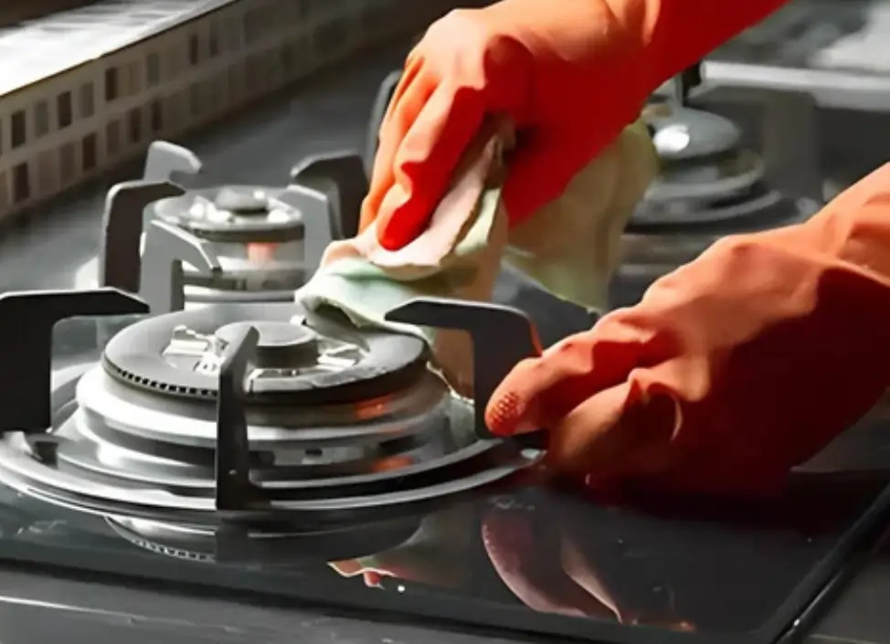 Person wearing orange gloves cleaning a gas stove burner with a cloth on a glass-top stove surface.
