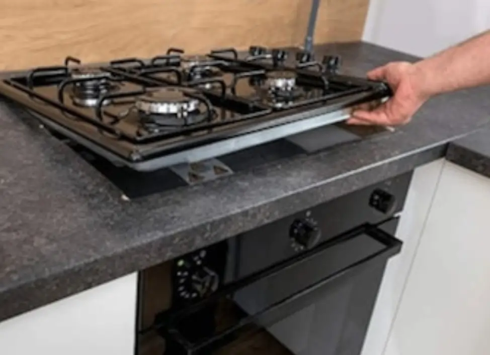A person installing a four-burner gas stove into a kitchen countertop above a built-in oven.