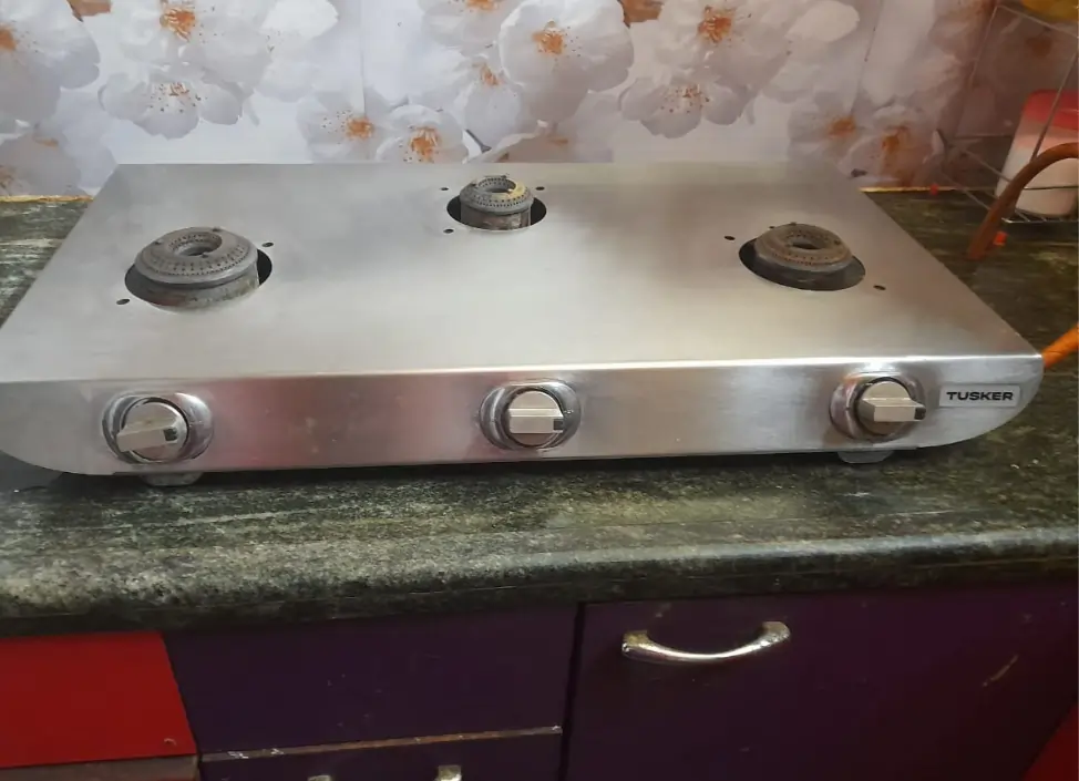 A three-burner stainless steel gas stove placed on a kitchen countertop with floral-patterned backsplash.